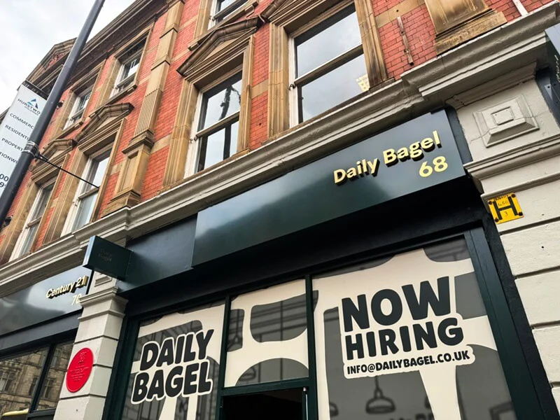 An image of the exterior signage for Daily Bagel, which also shows "Now Hiring" signs in the window.
